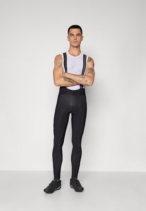 Schwarze Fahrradhosen mit verstellbaren Trägern und gepolsterter Sitzfläche. Unter einem weißen Tanktop getragen. Glatte Textur, taillierte Passform und minimalistische Details.
