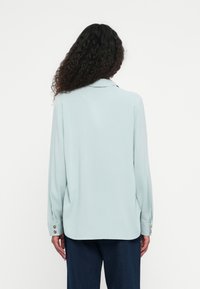 Jasnoniebieska bluzka z długimi rękawami i kołnierzykiem. Posiada złote guziki na mankietach. Gładki, lejący materiał z luźnym krojem.