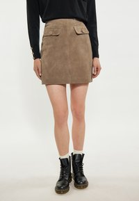Tan mockaskjorts med två framfickor och dekorativa knappar, bärs med svarta snörstövlar och vita ankelstrumpor.