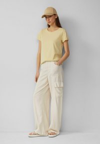 Lichtgele t-shirt met korte mouwen, crèmekleurige cargobroek met wijde pijpen en zijzakken, beige pet en slippers op een neutrale achtergrond.