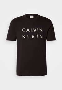 T-shirt en coton noir avec l'inscription "CALVIN KLEIN" en lettres métalliques argentées en gras sur la poitrine. Col rond classique et manches courtes.