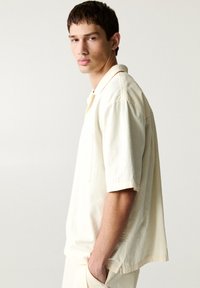 La camicia a maniche corte color crema presenta una vestibilità rilassata, un tessuto texturizzato, un colletto a punta e una singola tasca sul petto.