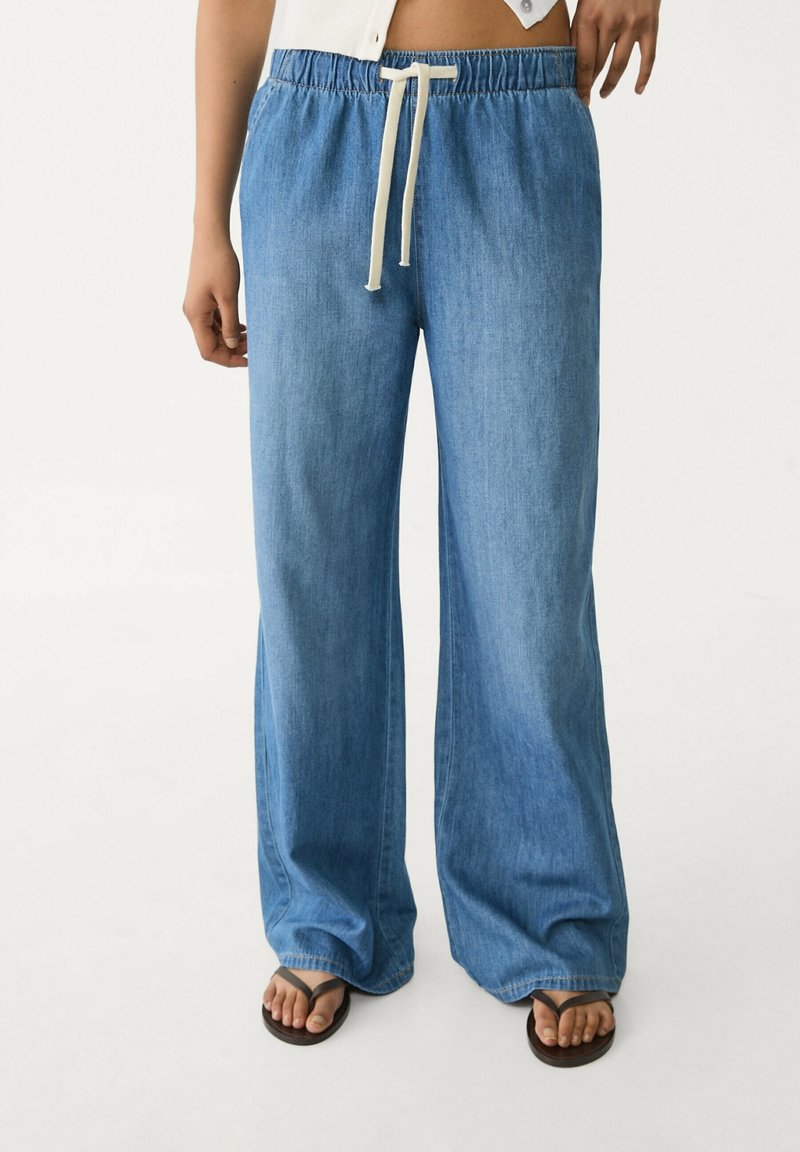 Kvinna som bär ljusblå vida jeansbyxor med elastisk midja och vit dragsko, i kombination med svarta flip-flops och en kort topp.