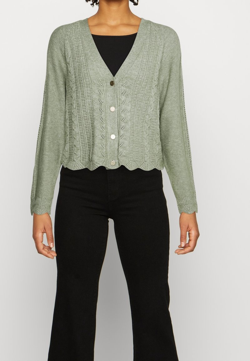 Femme portant un cardigan vert clair en tricot avec un motif de feuilles et des bords festonnés, associé à un haut noir et un pantalon noir.