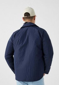 Chaqueta ligera de color azul marino con un acabado texturizado, de mangas largas y cuello clásico, combinada con jeans de mezclilla. Se lleva una gorra blanca.