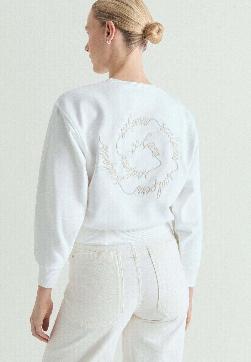 Mujer con cabello rubio recogido en un moño, que lleva pantalones blancos y una sudadera blanca con bordado en cursiva en la espalda en forma circular.