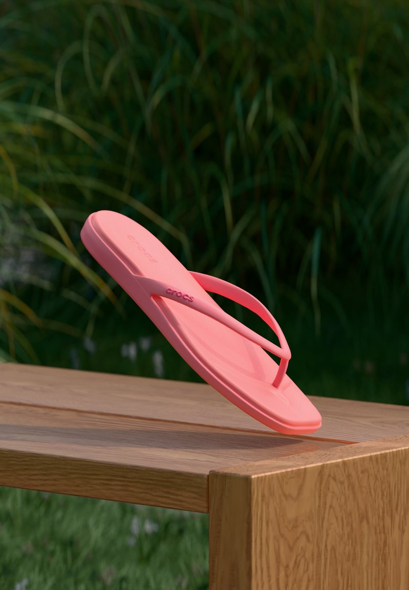 Ροζ κοραλλί σαγιονάρα Crocs ισορροπημένη στην άκρη ενός ξύλινου πάγκου με πράσινο φύλλωμα στο φόντο.