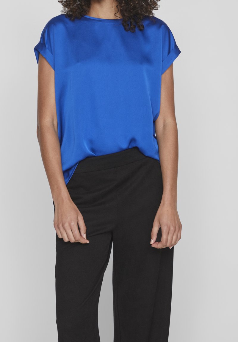 Femme portant un chemisier bleu vif à manches courtes, rentré dans un pantalon noir taille haute, debout devant un fond uni.