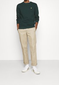 Sudadera verde de manga larga con un pequeño logo amarillo, chinos beige y zapatillas blancas. Texturas suaves con un ajuste casual.