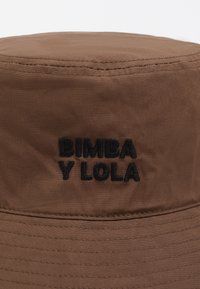 Sombrero de cubo marrón hecho de un tejido texturizado con un ala cosida. Presenta un logotipo bordado en negro que dice "BIMBA Y LOLA."