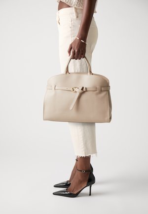 Beige leather handbag with a rounded top, a front strap and bow detail, held by a person wearing white jeans and black heels.