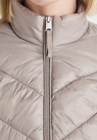 Light beige puffer jacket featuring a high collar, chevron stitching, and a front zipper with a silver pull tab. Soft, quilted texture.