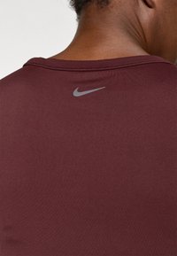Bordo sportska majica s glatkom teksturom. Ima sivi Nike logo na leđima, okrugli izrez i bez vidljivih uzoraka ili ukrasa.
