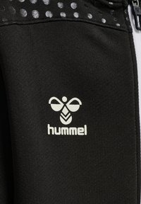 Giacca sportiva nera in tessuto con una texture liscia. Presenta un logo "hummel" bianco e motivi a pois argentati vicino alla spalla. Dettaglio della zip visibile.