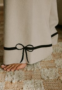 Beež pleed musta heegeldatud paelaga ääres, mis on visatud jalale tekstuurse kootud vaiba peale.