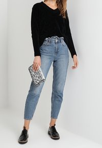 Černý svetr s výstřihem do V, světle vyprané džíny s vysokým pasem, lesklé černé boty, drží texturované stříbrno-černé psaníčko s hadím vzorem.