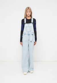 Lichtblauwe denim overall met een losse pasvorm, voorzien van een voorzak, verstelbare banden en een strikdetail bij de taille. Lange mouwen top eronder.