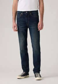 Bărbat îmbrăcat cu blugi slim-fit de culoare albastră închis, tricou alb și adidași negri cu talpă joasă și șireturi albe, stând în fața unui fundal simplu.