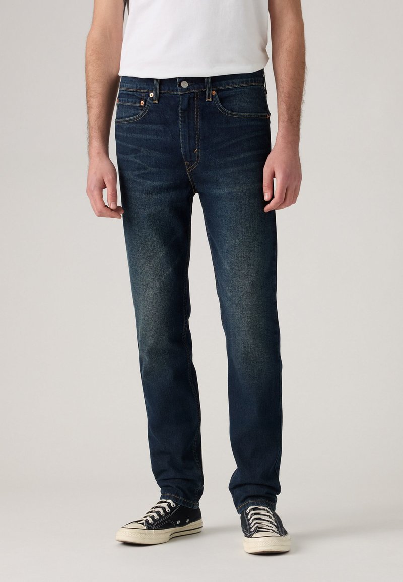 Bărbat îmbrăcat cu blugi slim-fit de culoare albastră închis, tricou alb și adidași negri cu talpă joasă și șireturi albe, stând în fața unui fundal simplu.