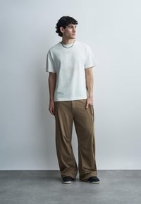 Νέος άνδρας με σγουρά σκουρόχρωμα μαλλιά, φοράει λευκό t-shirt, καφέ φαρδιά παντελόνια, μαύρα παπούτσια και ασημένια αλυσίδα, στέκεται μπροστά σε απλή τοίχο.