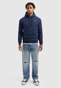 Man in een marineblauwe hoodie, lichtblauwe ripped jeans en zwart-witte sneakers, staand tegen een effen achtergrond.