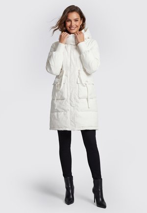 Femme souriante portant un long manteau blanc matelassé à capuche, un legging noir et des bottines noires à talons hauts, debout devant un fond uni.