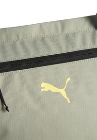 Bolsa de tela verde claro con una sutil textura estriada, que cuenta con un cierre negro y un logo de Puma amarillo en el frente.