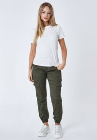 Camiseta blanca de manga corta con letras sutiles, combinada con pantalones cargo verde oliva que presentan múltiples bolsillos y puños elásticos. Zapatillas blancas.