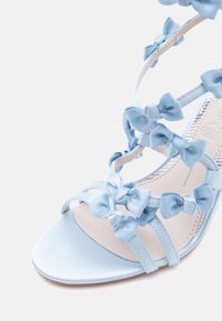 Ανοιχτό μπλε σανδάλι με ψηλό τακούνι και λουράκια, διακοσμημένο με πολλούς μικρούς φιόγκους κατά μήκος των λουριών και σχεδιασμό με ανοιχτά δάχτυλα.