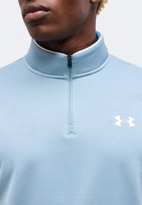 Άνδρας που φοράει ανοιχτό μπλε Under Armour pullover με φερμουάρ μέχρι τη μέση, υφασμάτινη υφή και λευκό λογότυπο στο στήθος, κοντινό πλάνο στον λαιμό και το πάνω μέρος του κορμού.