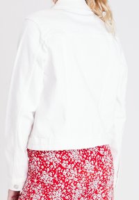 Weiße Jeansjacke mit klassischem Kragen, kurzem Schnitt und geknöpften Manschetten, kombiniert mit einem roten Blumenrock im Hintergrund.