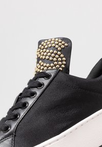 Sneaker nera con borchie dorate sul tallone in pelle nera, tomaia in tessuto testurizzato, lacci neri piatti e suola in gomma bianca.