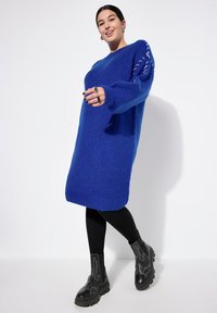Blå strikket sweaterkjole med lange ærmer, ribbede kanter og dekorativ stikning på ærmerne. Bæret med sorte leggings og kraftige støvler.
