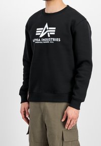Felpa nera in misto cotone con scollo rotondo, caratterizzata da un logo grafico bianco "Alpha Industries" e polsini e orlo a costine.