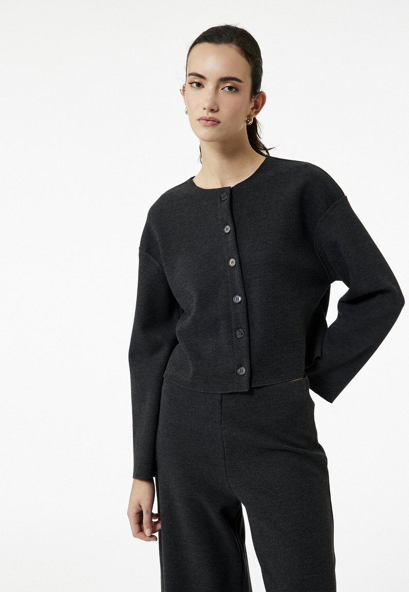 Cardigan corto nero con maniche lunghe, scollatura rotonda e chiusura frontale con bottoni. Morbido tessuto a maglia, abbinato a pantaloni coordinati.