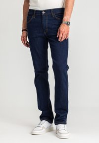 Ciemnoniebieskie dżinsy z denimu z pomarańczowym przeszyciem, pięciokieszeniowym krojem i prostymi nogawkami, noszone z białymi trampkami.