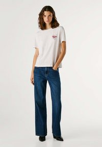 Femme aux cheveux mi-longs portant un T-shirt graphique blanc, un jean large bleu et des chaussures noires à bouts pointus, debout avec une main dans la poche.