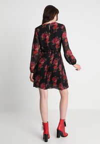 Mustassa kukallinen pitkähihaisen mekon, jossa on laskostettu hame, pinkki- ja punakukkaistekstejä; aukko selkäpuolella, yhdistettynä punaisiin korkokenkiin.