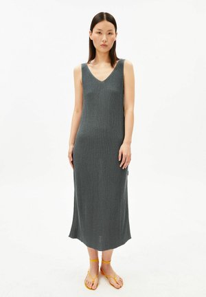 Femme debout portant une robe midi en maille côtelée grise sans manches et des sandales plates jaunes à brides sur un fond blanc uni.