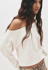 Weißes sweatshirt mit schulterfreien Ärmeln aus weichem Stoff. Verfügt über lange Ärmel, einen cropped Saum und eine zarte silberne Halskette.