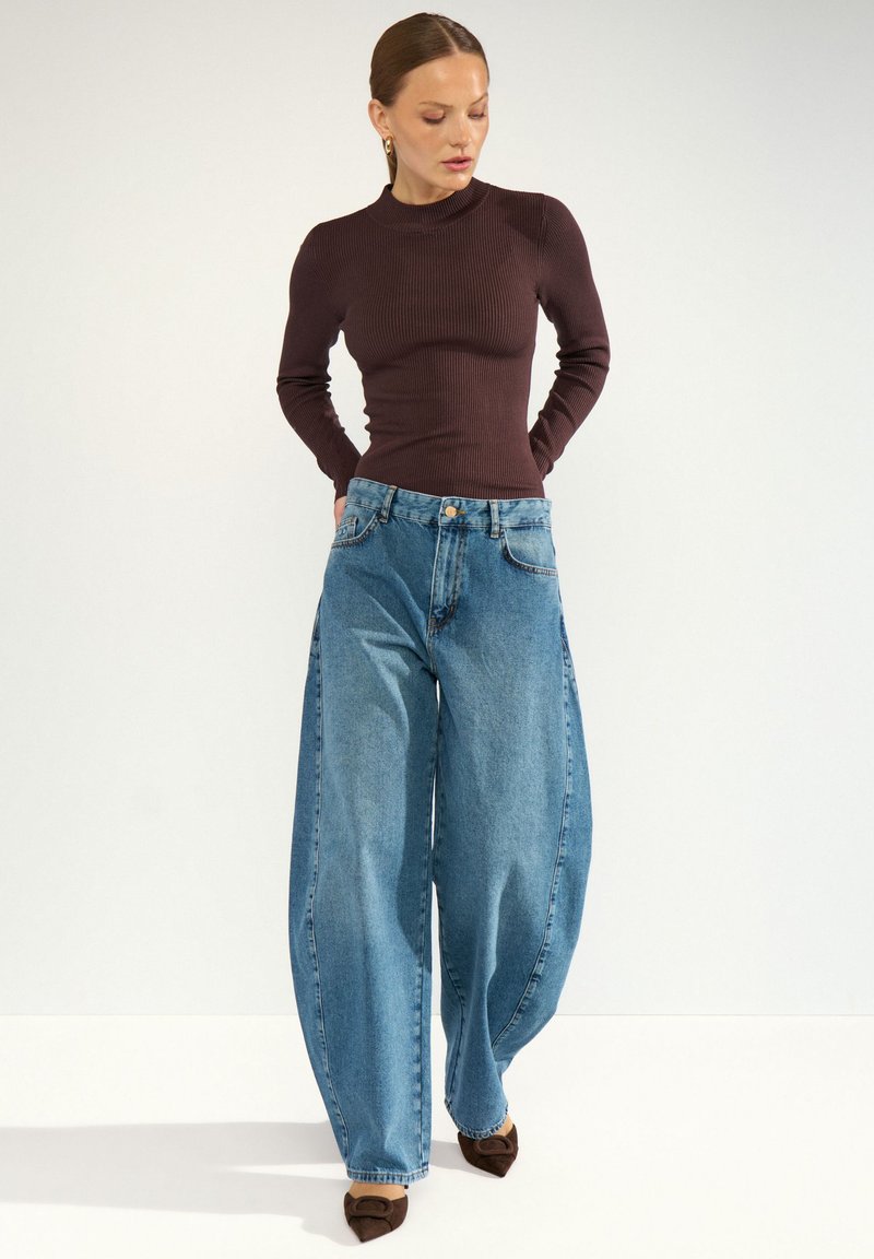 Femme portant un haut à manches longues foncé, un jean bleu à jambes larges et des chaussures pointues marron, debout les mains dans les poches arrière sur fond blanc.