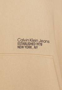 Felpa beige realizzata in materiale morbido con tasca frontale. Testo nero ricamato: "Calvin Klein Jeans FONDATO NEL 1978 NEW YORK, NY."