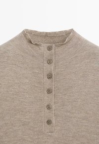 Beżowa koszulka henley z prążkowanym kołnierzykiem, pięcioma brązowymi guzikami z przodu oraz miękkim, teksturowanym materiałem.