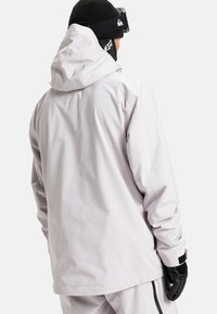 Person, der eine weiße Kapuzenjacke, eine schwarze Strickmütze, eine Skibrille und schwarze Handschuhe trägt, sieht mit dem Rücken zu einem schlichten weißen Hintergrund.