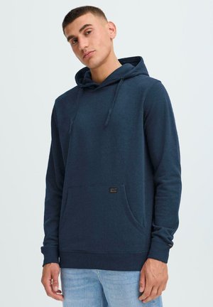 Marineblå hettegenser i myk stoff, med en frontlomme, snorhette, og en liten merkeetikett nær lommen. Jevn tekstur, avslappet design.