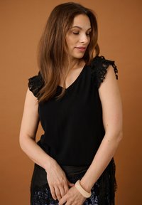 Femme aux longs cheveux bruns portant un haut noir à manches en dentelle et un bracelet beige, regardant vers le bas sur un fond marron uni.