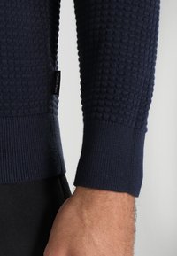 Mörkblå stickad tröja med en strukturerad mönster, ribbade muddar och en Calvin Klein-label på sidan. Närbild av ärm och tygdetalj.