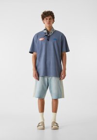 Blå overdimensjonert T-skjorte med grafiske trykk, falmede denimshorts, beige slip-on sko og lys gule sokker. En enkel bakgrunn.