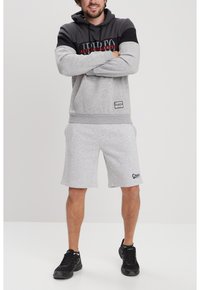 Sudadera gris con detalles en negro, con un logo en la parte delantera. Combinada con pantalones cortos grises y zapatillas negras. Look deportivo casual.
