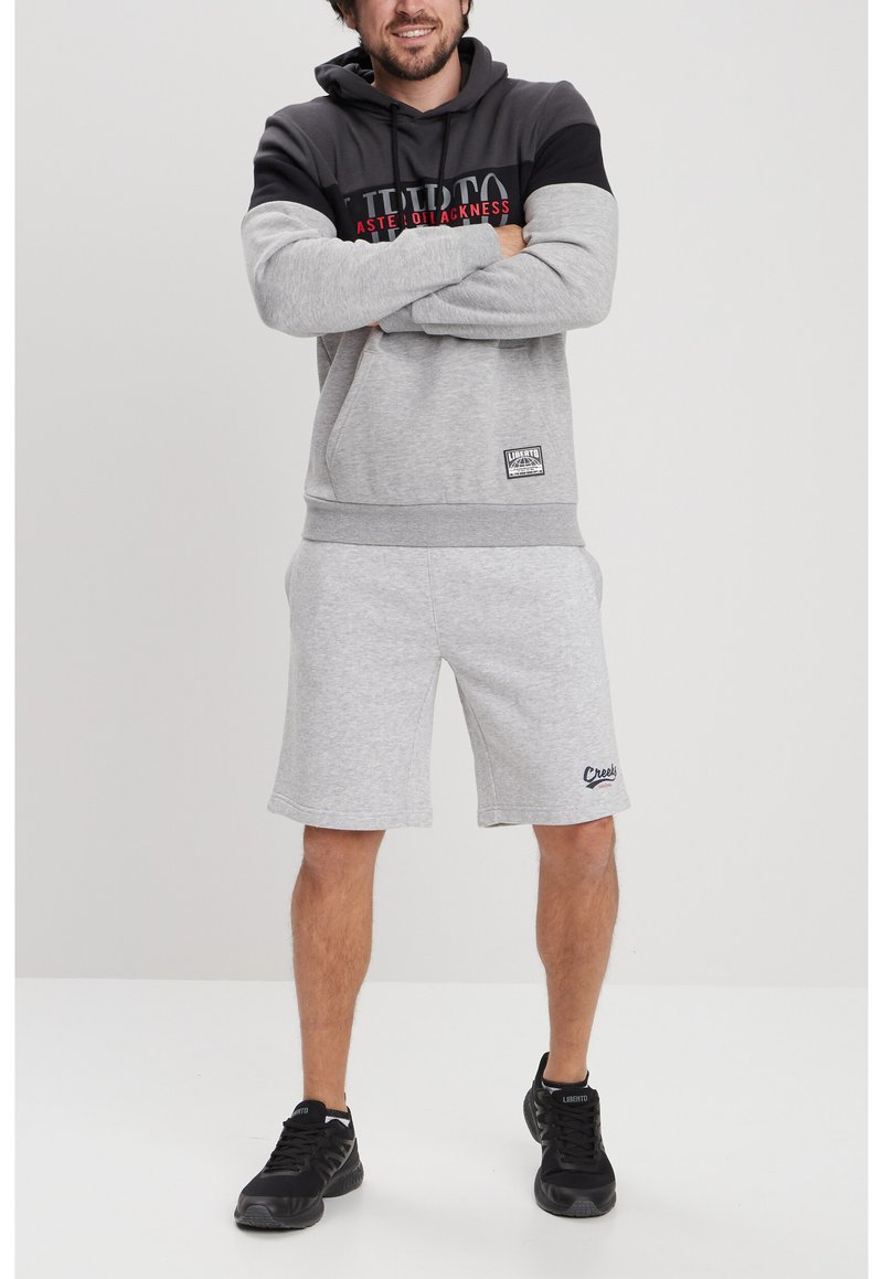 Sudadera gris con detalles en negro, con un logo en la parte delantera. Combinada con pantalones cortos grises y zapatillas negras. Look deportivo casual.